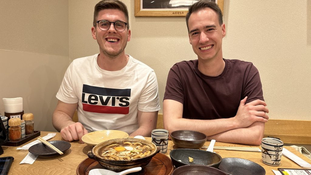 Two young white men smile for the camera sitting in a table booth with bowls of misonikomi udon and hitsumabushi eel before them.