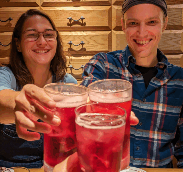 A younger man and woman make a toast with the cameraperson, holding up glasses of a fizzy red drink.