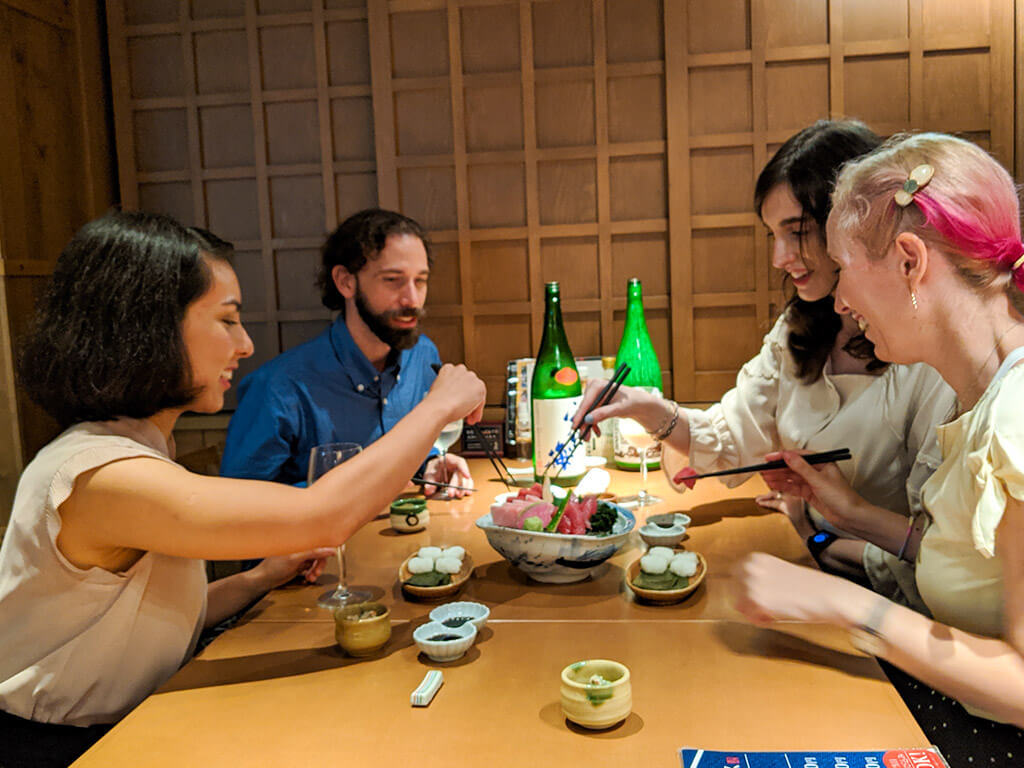 Three guests and our CEO, Elly, smiling and toasting with sake cups on our Nagoya Sake Tasting Night Tour. Four green sake bottles and several plates of sashimi are scattered across the table.