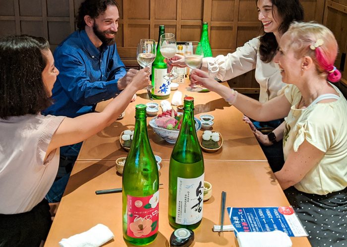 Three guests and our CEO, Elly, smiling and toasting with sake cups on our Nagoya Sake Tasting Night Tour. Four green sake bottles and several plates of sashimi are scattered across the table.