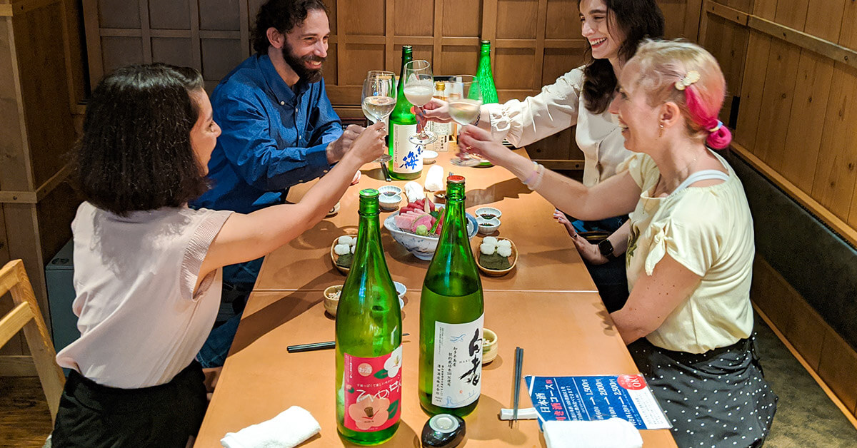 Three guests and our CEO, Elly, smiling and toasting with sake cups on our Nagoya Sake Tasting Night Tour. Four green sake bottles and several plates of sashimi are scattered across the table.