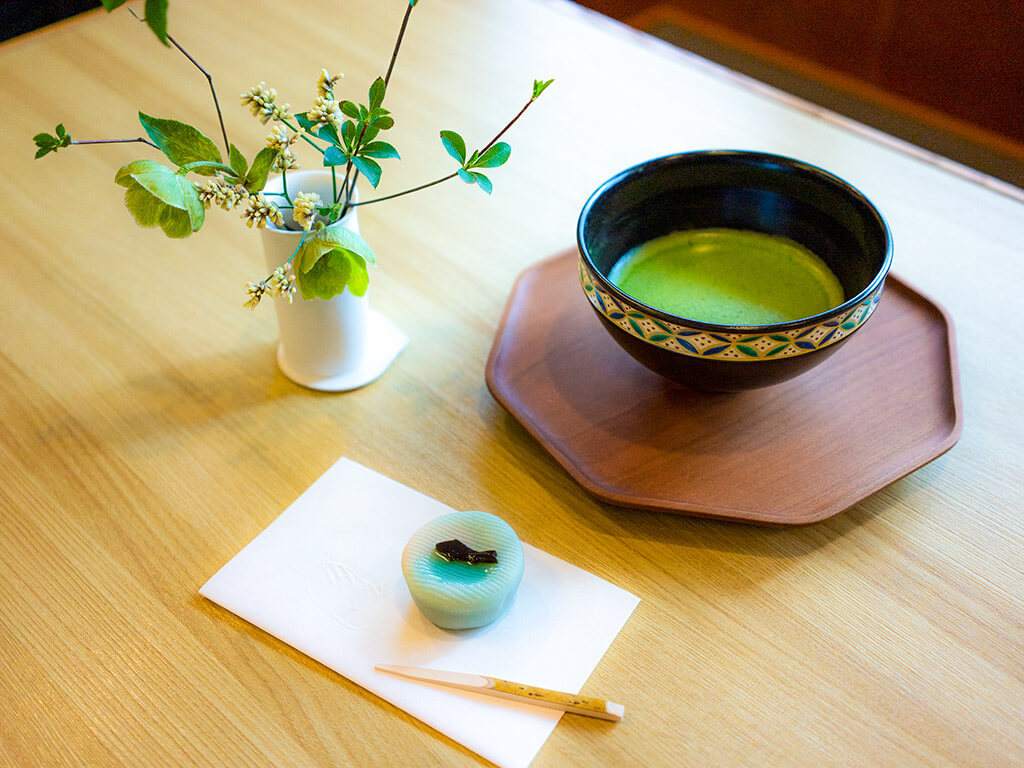 A small vase with a few white flowers poking out, a turquoise wagashi sweet with a black fish on top and a skewer, and a bowl of matcha on a wooden hexagonal plate, all on a light wood table.