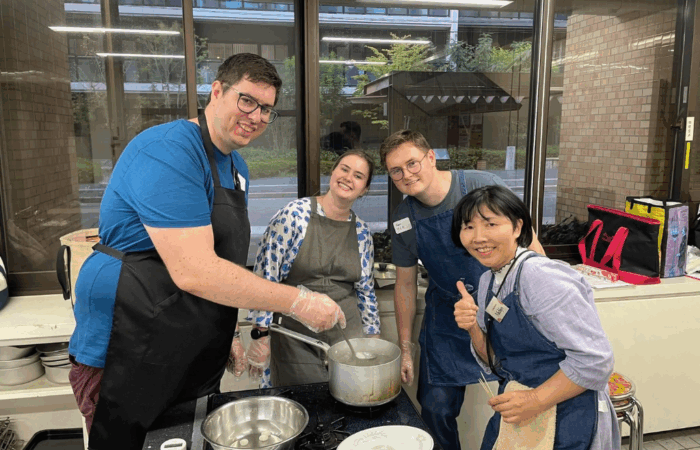 Three students wearing aprons pose with instructor Junko over a set of pots making dashi.