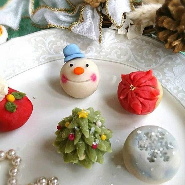 Nerikiri Five christmas-themed nerikiri sitting on a white plate. Clockwise from top: a snowman, a poinsetta flower, a silver snowflake, a wreath, and Santa.