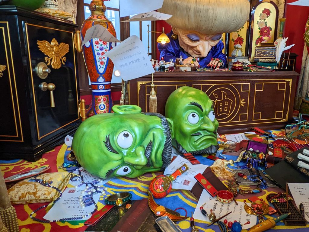 Two green heads sit atop some clutter as Yubaba from "Spirited Away" (a woman with purple eyeshadow, a pointed nose, and a large head of white hair) works at her desk.
