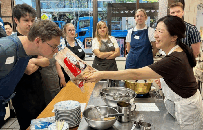 Instructor Junko holds out a bag of Katsuo bonito flakes for a student standing with a group around a kitchen island in an industrial kitchen.