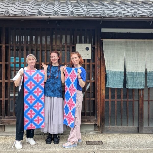 Arimatsu Shibori Experience An artisan and two participants hold up their red and blue indigo designs in front of a traditional Japanese shop.