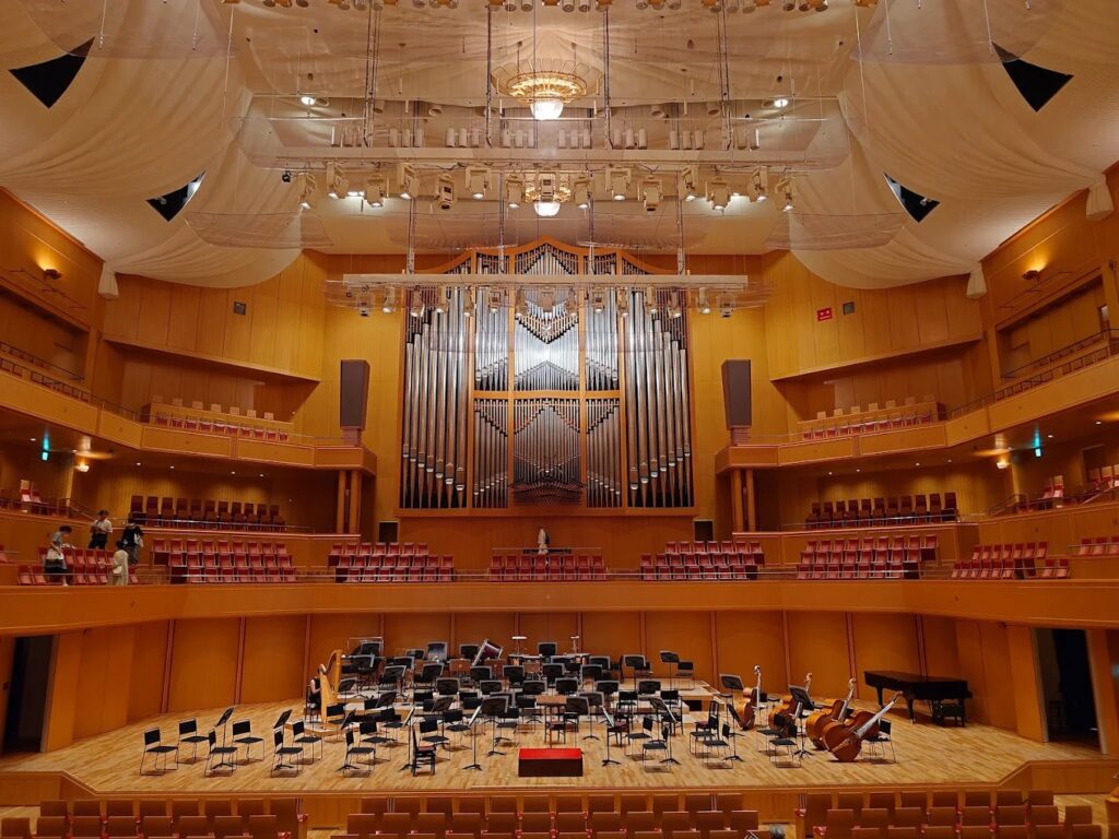 The dramatic stage of Aichi Arts Center, with red chairs in rows above the stage, an organ-inspired bay window, and chairs and larger instruments set up on stage.
