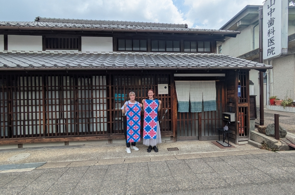 The arimatsu shibori artisan and a participant showing off their red and blue indigo designs in front of a traditional Japanese storefront.