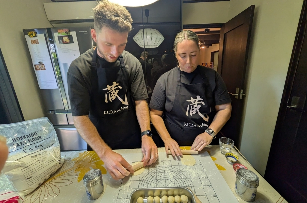 Two participants rolling out gyoza dough in the kitchen, wearing black aprons reading "Kura cooking" in English and Japanese.