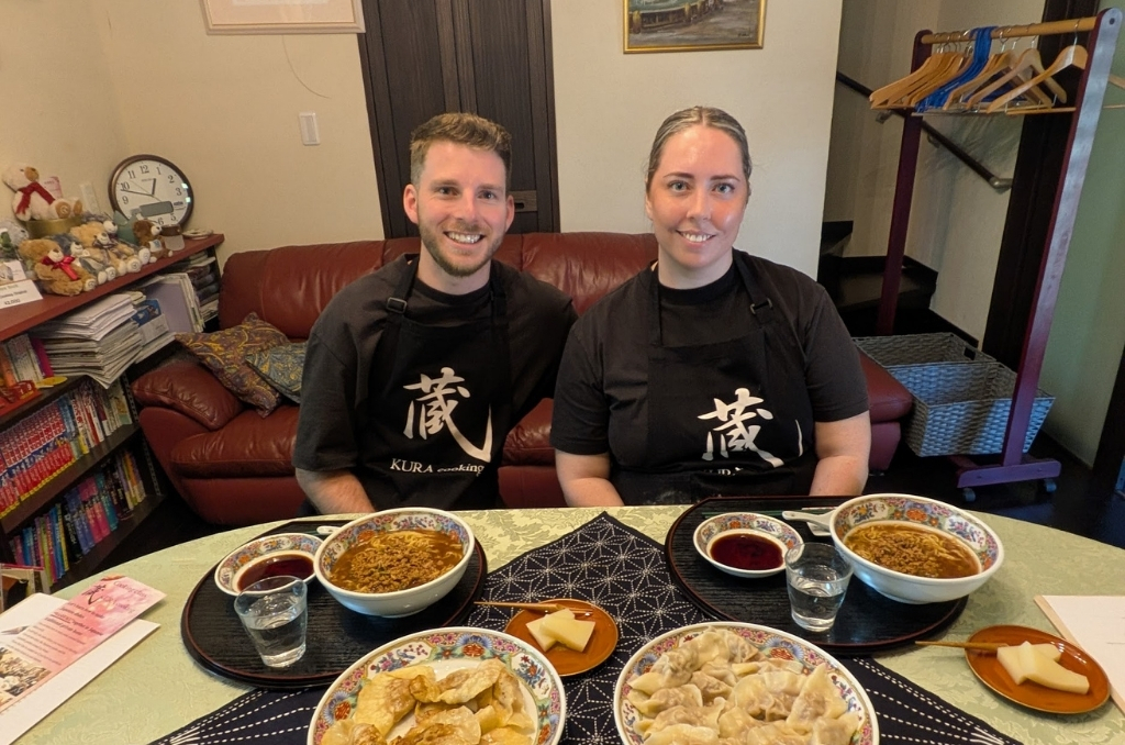 A man and woman wearing black aprons sit down before a full table of homemade ramen and gyoza dishes.