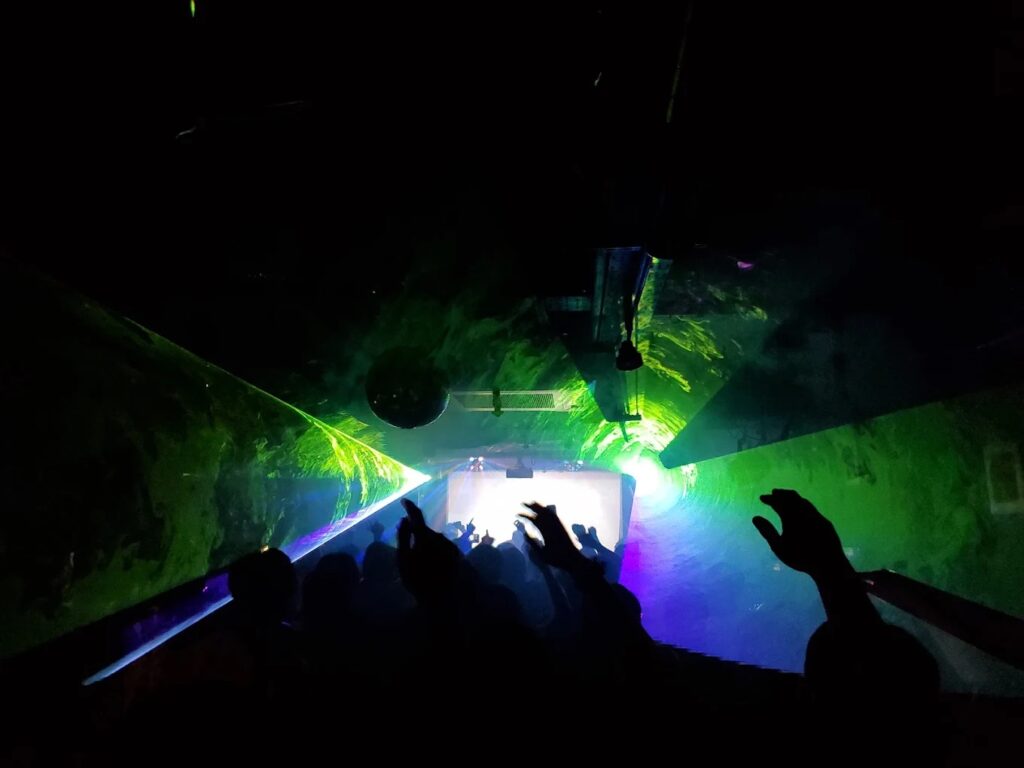Inside the club at JB's, with the crowd's hands raised and a swirl of green, purple, and blue lights projecting in the space.
