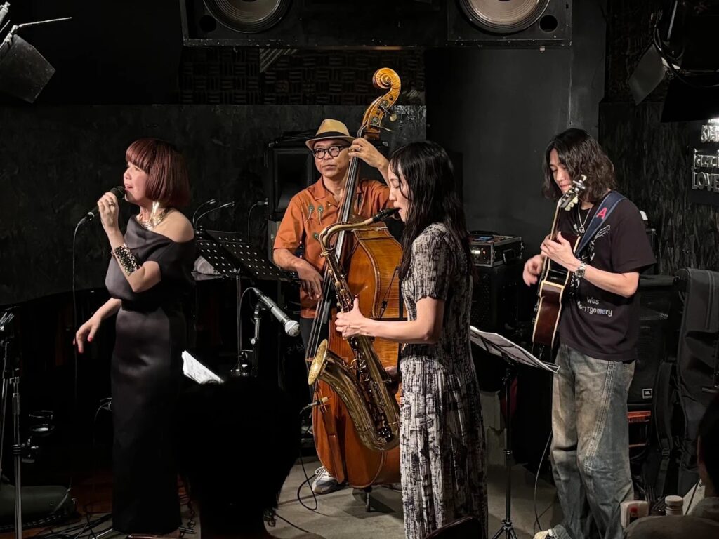 Four performers at Jazz Inn Lovely: a woman singing, a woman playing saxophone, a man playing the cello, and a man playing acoustic guitar.