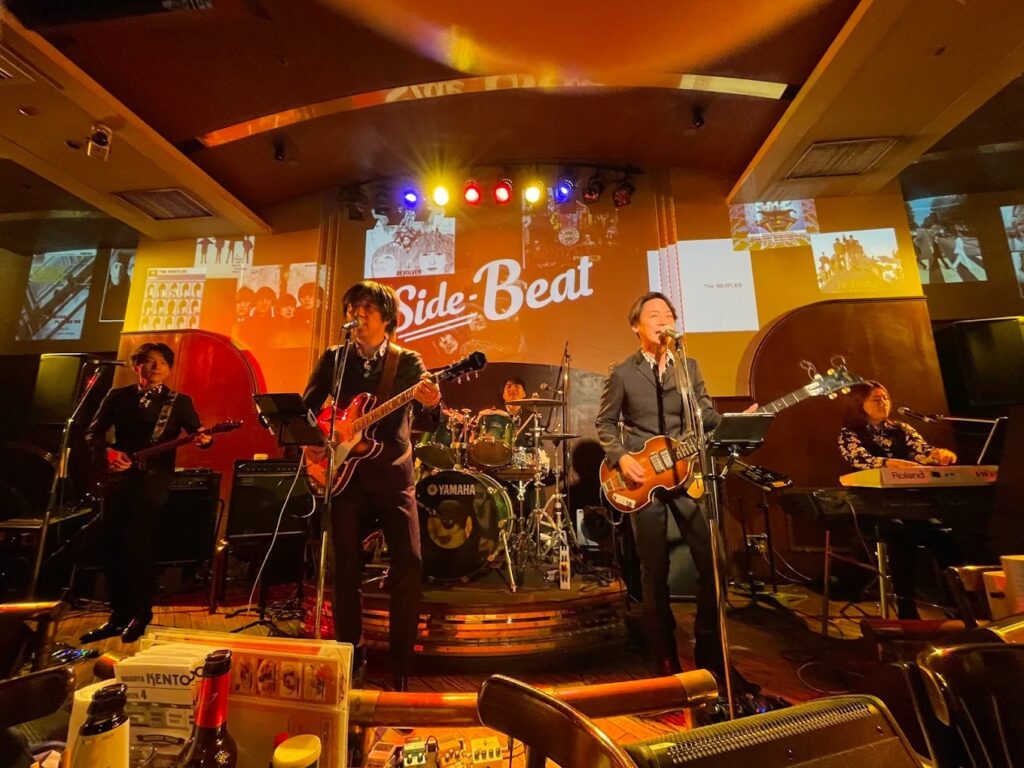 Performers on stage at Kento's Bar: two male singers playing electric guitars, a man playing the bass, a man on the drums, and a woman playing the keyboard in front of a warm yellow and wood background.