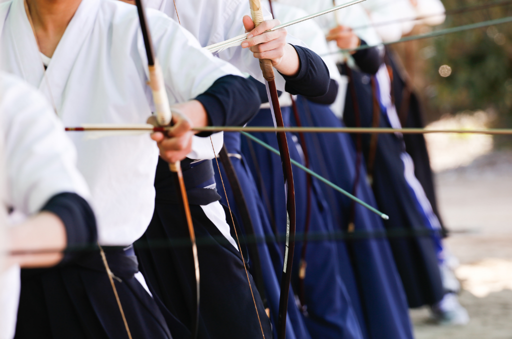 A line of Kyudo archers in white and navy hakama drawing their bows, with only torsos and arms visible in the frame.
