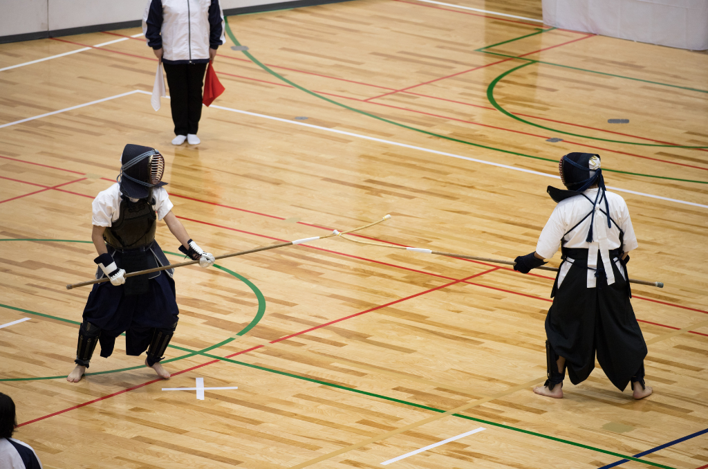 Two naginata swordsmen engaged in a duel in a school gym with two coaches/referees standing off to the side on opposite corners, holding a white and red flag in each hand.