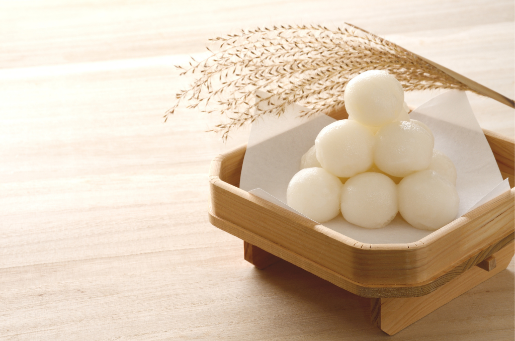 A pyramid of tsukimi dango on a wooden square plate with a branch of Japanese pampas grass draped behind.