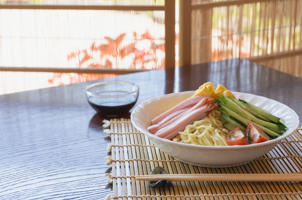 A bowl of hiyashichuka soba, with the yellow soba noodles on the bottom, thin, long slices of ham, cucumber, rolled egg, and tomatoes on top. The white bowl is sitting on a mini tatami-style placemat, with chopsticks resting in front and a clear, small bowl of soy sauce on the lefthand side in front. In front of the table, a window gives a partial view of a red, leafy bush in the sunlight.