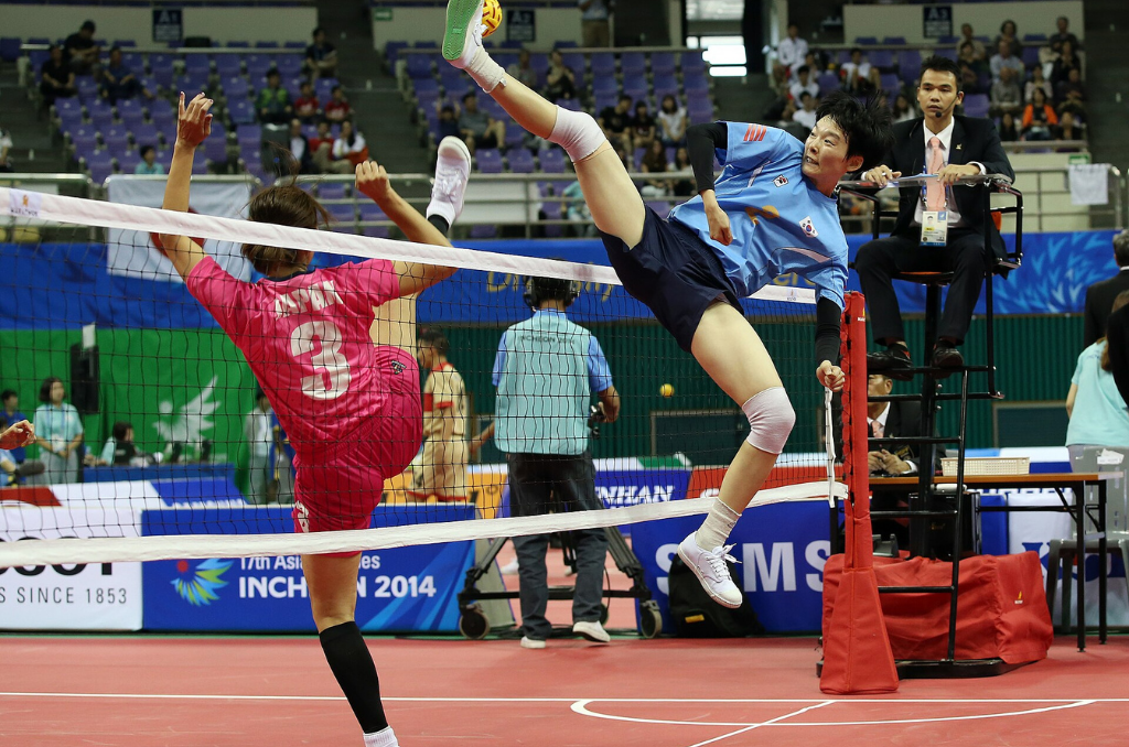 Two Sepak Takraw players on opposite sides of the net in burgundy and blue uniforms kick their right legs and fly into the air to reach a small yellow and red ball while a coach in a suit and tie looks on from a black high chair with a crowd in blue chairs behind him.