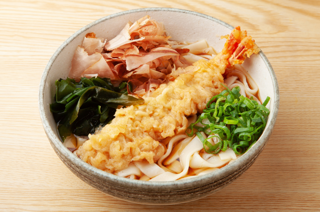 An eggshell bowl of hiyashi kishimen, with bonito flakes, seaweed, green onion, a large shrimp tempura, and wide kishimen noodles in a light brown broth.