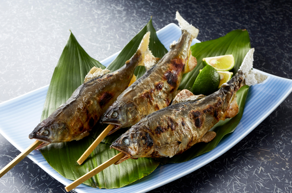 Three grilled ayu sweetfish on skewers rested on a plate topped with leaves and lemon slices.