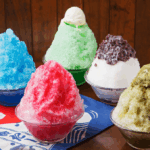 Five bowls of kakigori. Clockwise from middle: matcha with vanilla ice cream on top, vanilla with red bean paste on top, pistachio with pistachio ice cream on top, strawberry or raspberry, and ramune soda, all sitting on a blue, red, and white kakigori banner.