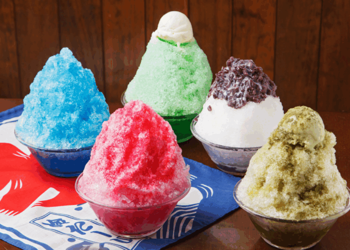 Five bowls of kakigori. Clockwise from middle: matcha with vanilla ice cream on top, vanilla with red bean paste on top, pistachio with pistachio ice cream on top, strawberry or raspberry, and ramune soda, all sitting on a blue, red, and white kakigori banner.