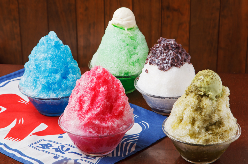 Five bowls of kakigori. Clockwise from middle: matcha with vanilla ice cream on top, vanilla with red bean paste on top, pistachio with pistachio ice cream on top, strawberry or raspberry, and ramune soda, all sitting on a blue, red, and white kakigori banner.
