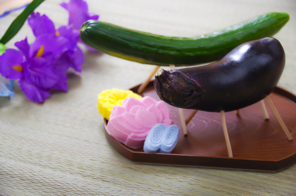 A cucumber and an eggplant propped up on four skewers on an oblong brown plate with lotus flowers and purple flowers arranged around it.