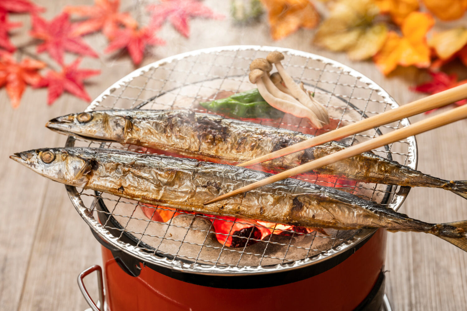 Two sanma fish being grilled on charcoal foil over a pot with autumn leaf decorations on the table in the background.