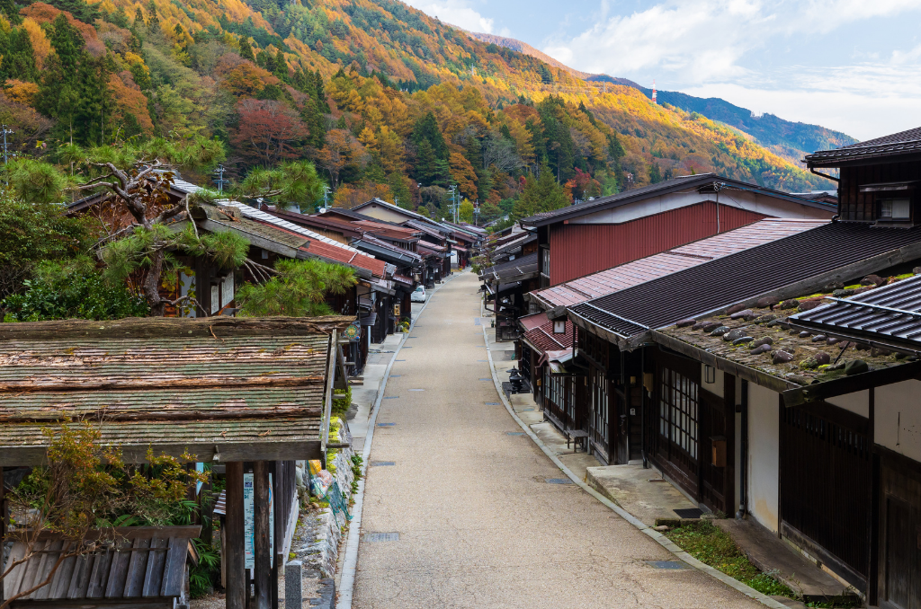 A wide-lens view of the Nakasendo walking path through traditional houses, with green, orange, and yellow fall colors in full bloom on the mountain in the background.