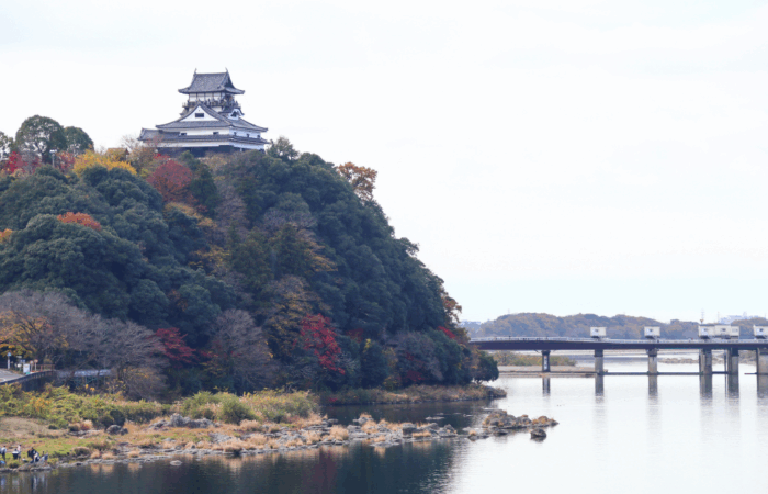 Inuyama Castle from a distance on a small mountain of trees in full fall colors of green, red, and orange, with a small bridge crossing the moat surrounding the castle.