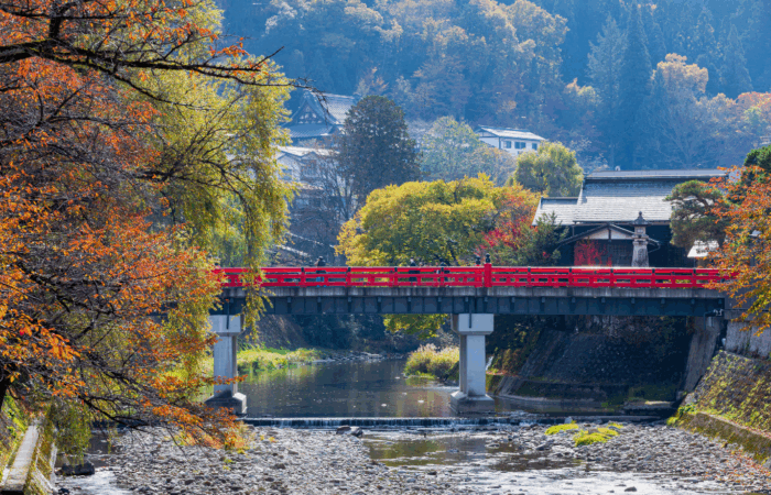 The red bridge of Takayama from a distance, running over a river lined with trees in full autumn colors.