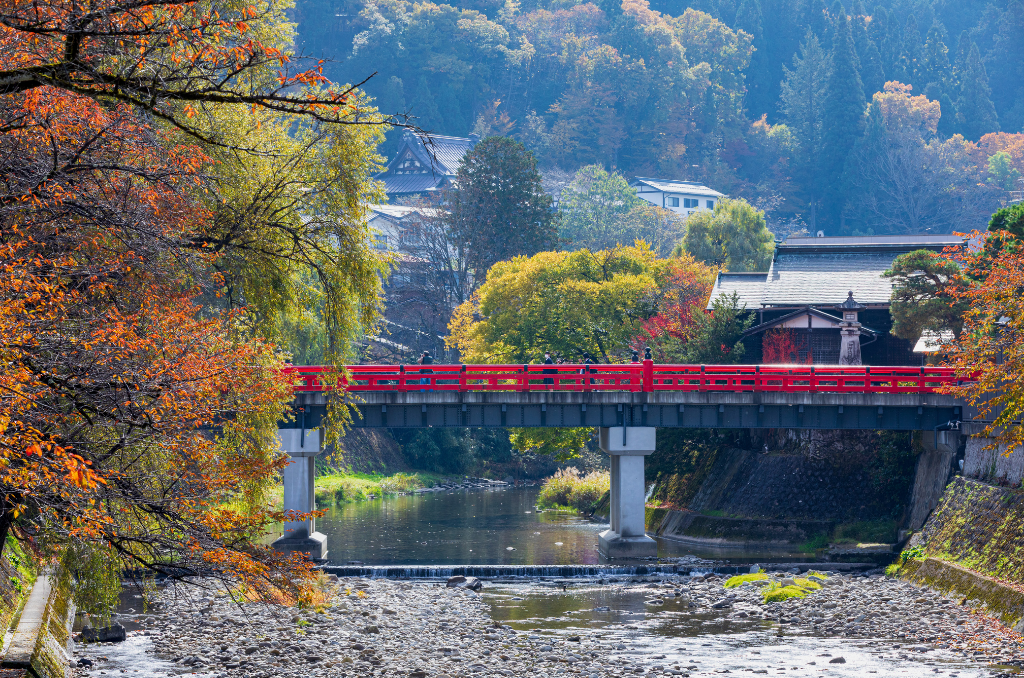 The red bridge of Takayama from a distance, running over a river lined with trees in full autumn colors.