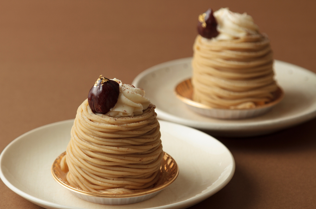 Two mont blanc chestnut desserts on gold foils on top of white plates in front of a tan background.