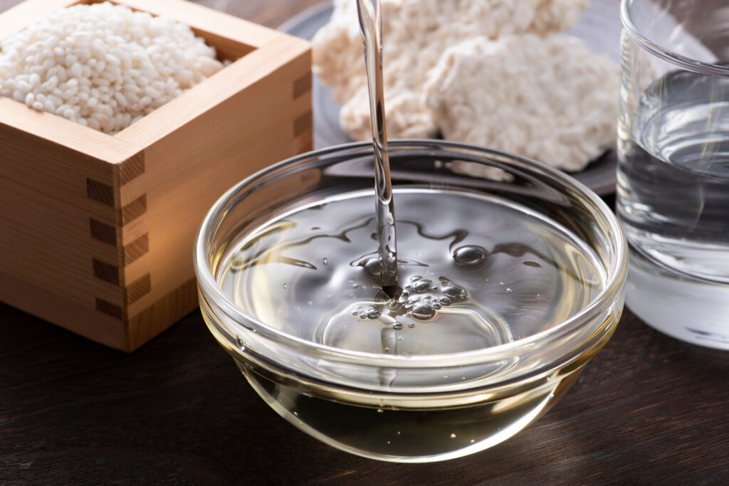 Mirin being poured into a small glass bowl, sitting in front of a wooden box of rice and a plate of koji mold.