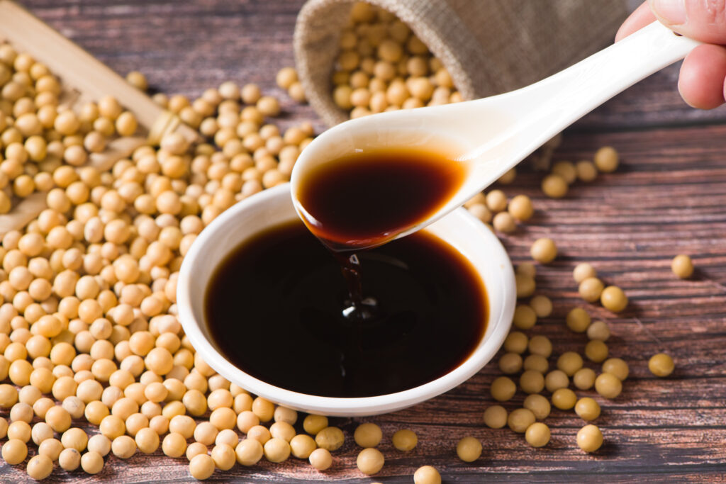 A white ladle is used to scoop up tamari in a small white dish, surrounded by soy beans spilling out onto the wooden table.