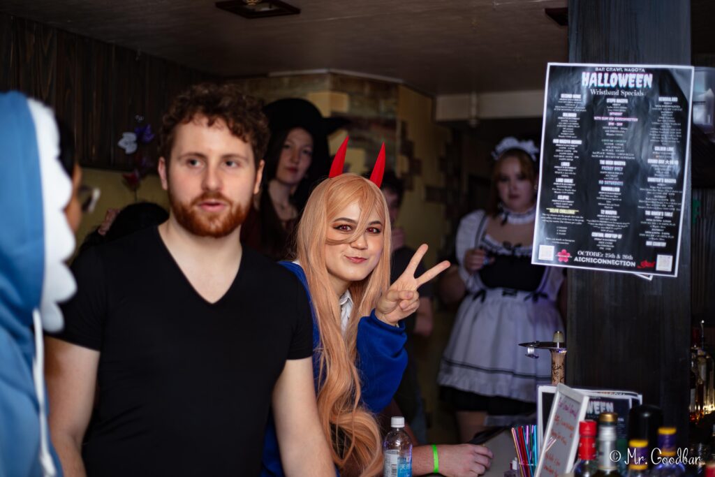 Participants pose in their Halloween costumes beside an event poster.